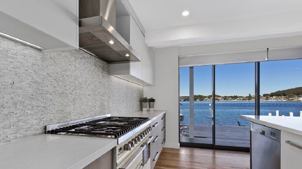 Modern coastal kitchen design with light cabinetry, stone backsplash, and water views through full-height glass doors—an example of coastal vs hampton kitchen style in a relaxed Australian setting.