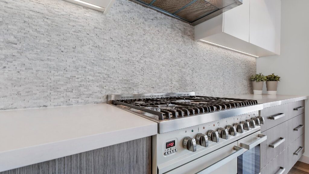 Close-up view of a modern coastal kitchen design in Australia, featuring a stainless steel gas cooktop, textured stone splashback, and sleek cabinetry with soft grey finishes.