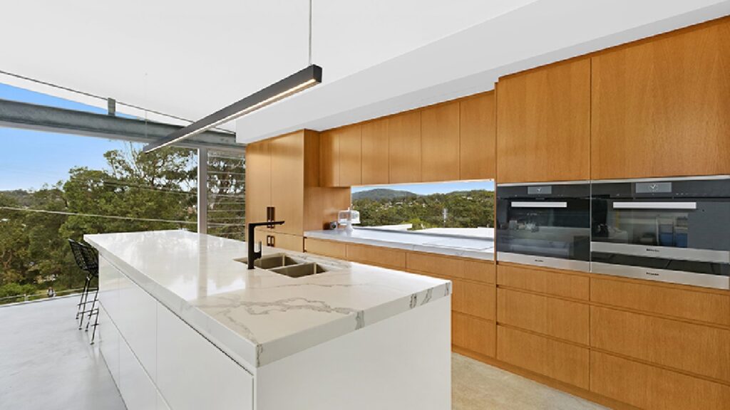 Modern coastal kitchen featuring warm timber cabinetry, a crisp white island bench with marble veining, and panoramic bush views—showcasing one of the 6 best colours for coastal kitchens.