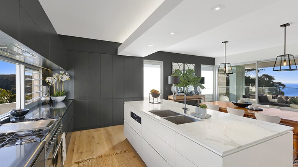 Modern open-plan kitchen with ocean views, featuring a white marble island, matte black cabinetry, natural timber flooring, and indoor greenery—an ideal example of how to design a coastal kitchen with elegance and functionality.